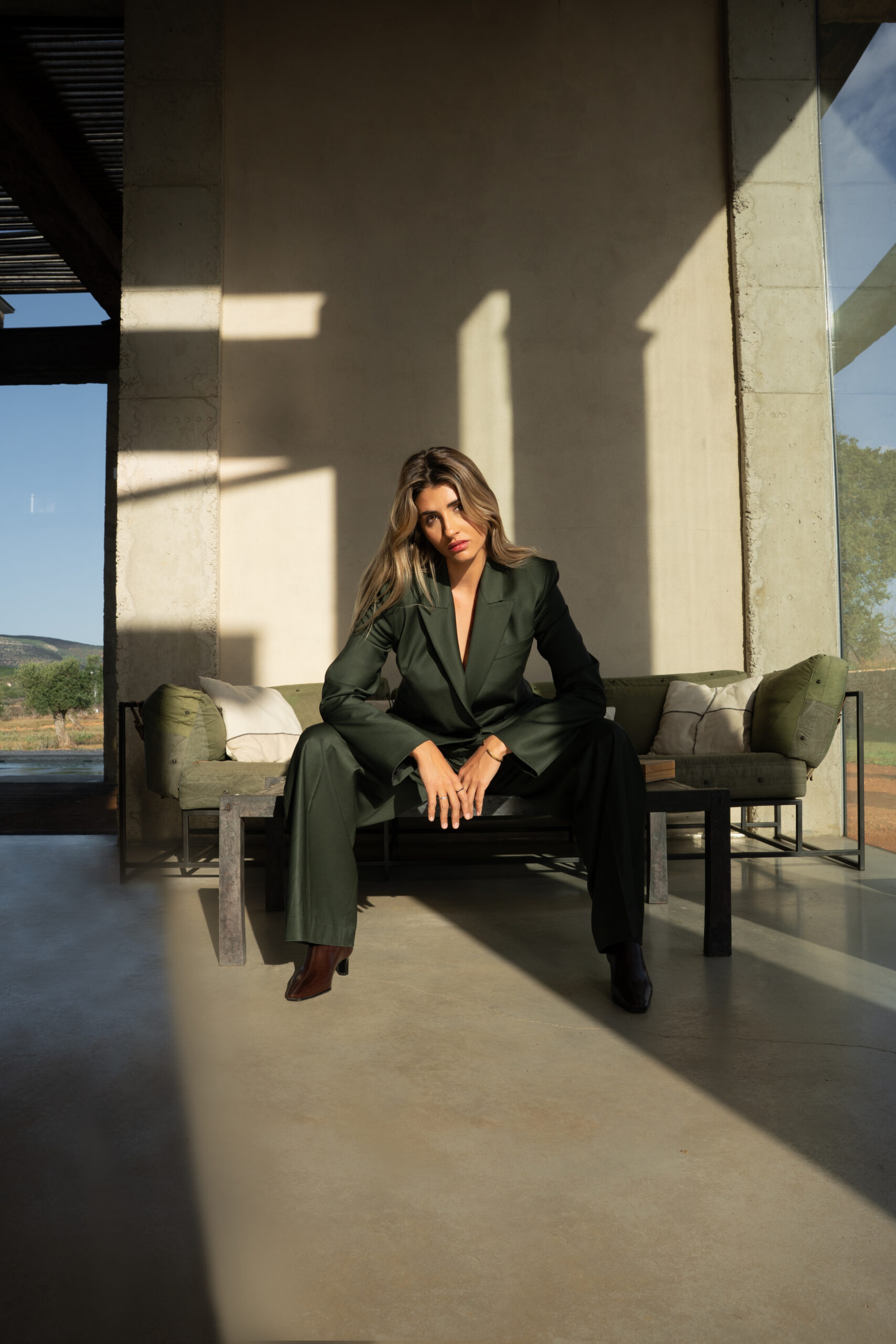 Woman in a dark green tailored suit seated on a bench under a concrete porch, countryside beyond
