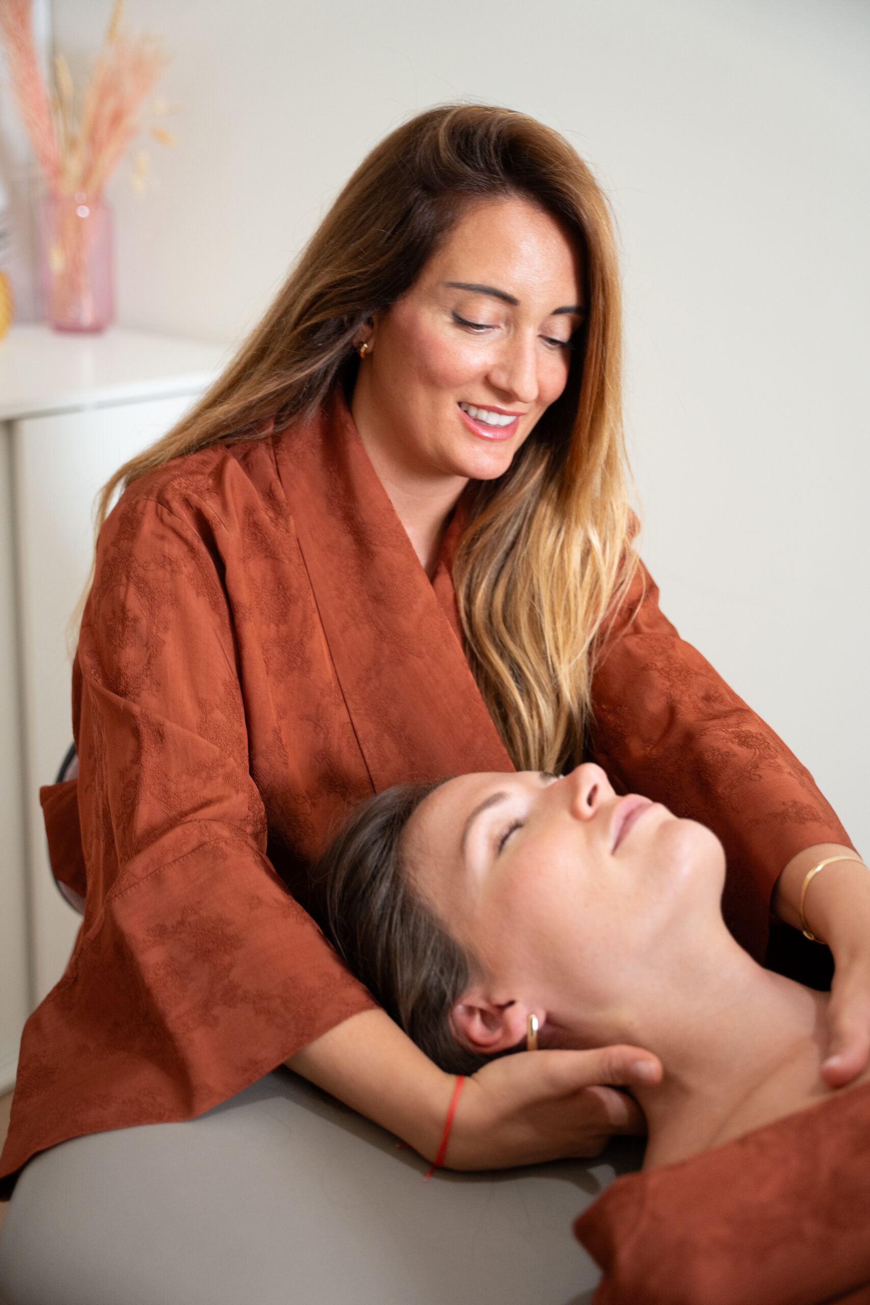 Two women in terracotta robes, one lying down receiving a head massage